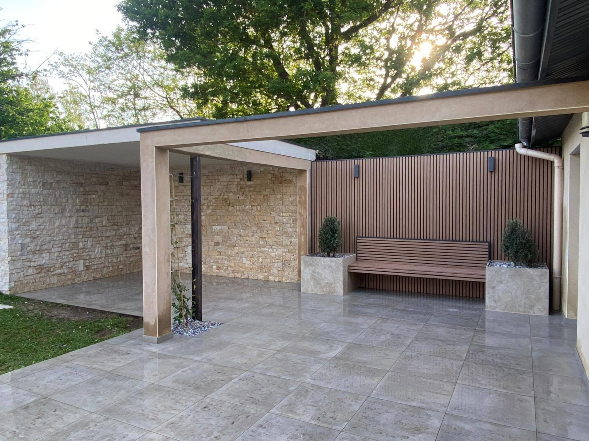 Terrasse extérieure en carrelage avec le mur du fond en bardage composite imitation clair-voie noir et bois clair avec un banc assorti placé entre deux pots de fleurs à Frontenas